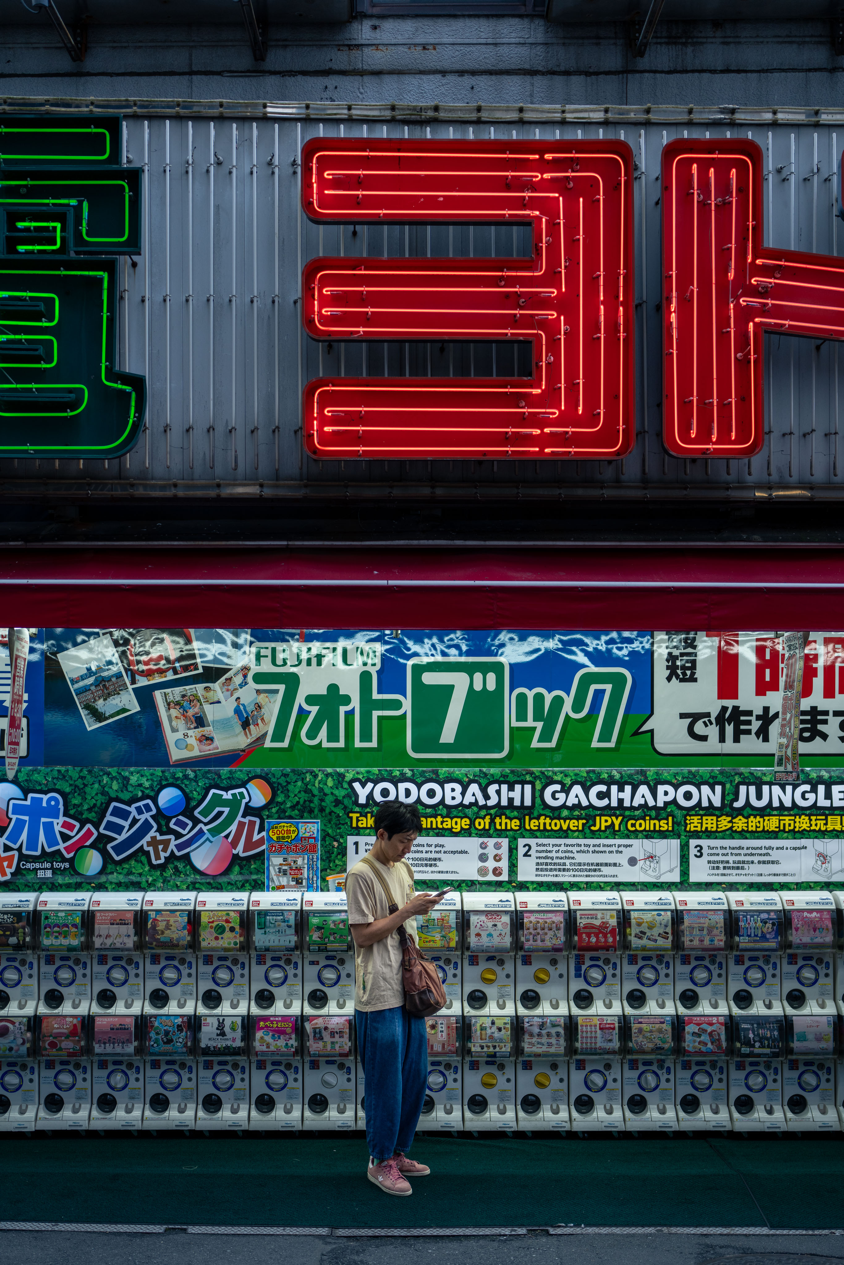A vertical photograph of a Tokyo street: floor-to-ceiling neon Japanese signage in red and green at the top, below it a printed wall of cartoon-bordered Yodobashi Gachapon Jungle ads, and at street level a bank of clear plastic capsule-toy vending machines. One man in a beige shirt stands quietly in front of them looking at his phone.