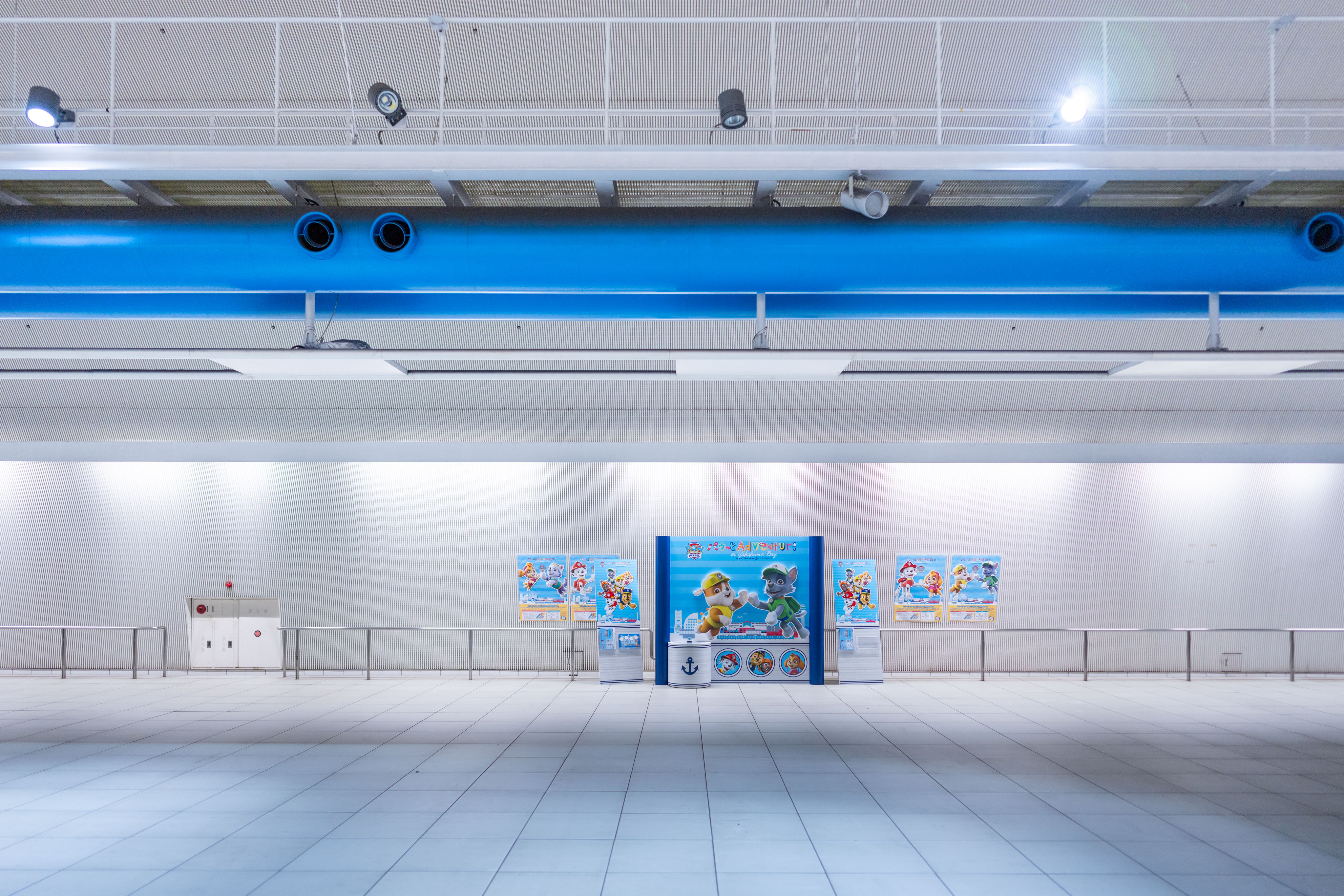An enormous, almost empty white-tiled metro concourse in Tokyo. Spotlights on a high ceiling pick out a bright blue overhead duct running the length of the hall. In the centre stands a small Paw Patrol promotional kiosk, its bright cartoon graphics dwarfed by the scale of the architecture around it.