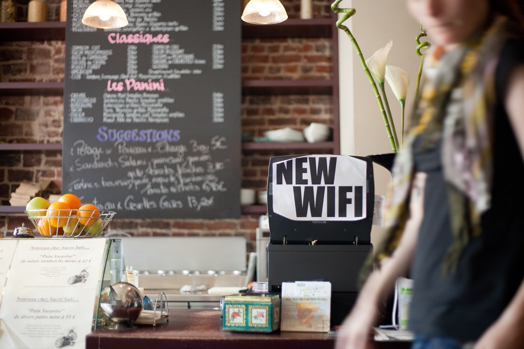 The counter of a French café in soft daylight. Behind it, a chalkboard menu with hand-lettered Classiques, Les Panini and Suggestions sections. To one side, a wire basket of oranges and lemons; to the other, a calla lily and the blurred shoulder of a woman in a scarf. Taped to the front of the till, a printed A4 sign in plain capital letters reading NEW WIFI.