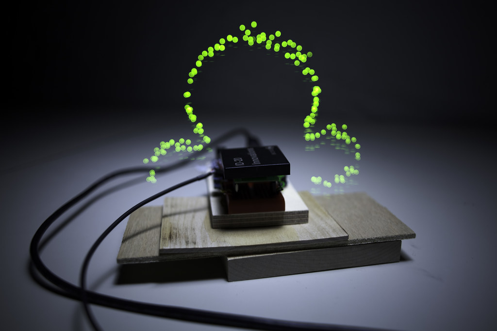 A small black RFID reader chip on a stack of pale wooden blocks. Floating above it in space, traced through long exposure, an arc of bright neon-green dots — the read-zone of the RFID antenna mapped as a 3D form by an LED probe waved through it.