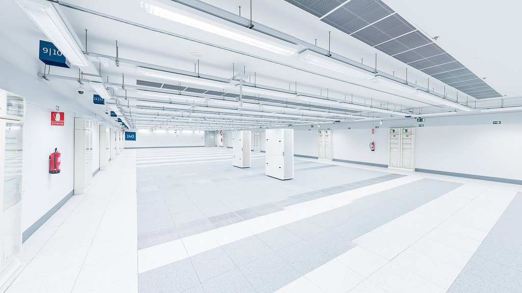 A stark white industrial hall inside a data centre. Polished tile floor, white plinths arranged in a grid, a long ribbon of fluorescent light running the length of the ceiling, blue numeric room signs (9 ten, eleven twelve, one-forty) hanging from the structure. Empty of people.