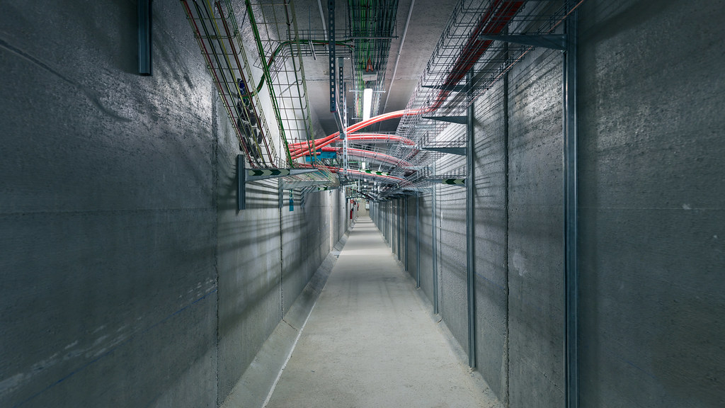 A long concrete utility tunnel inside the data centre. Bare grey walls and floor, vanishing into perspective. Overhead, dense cable trays carry bundles of red, orange and green data and power cables along the entire length of the corridor.