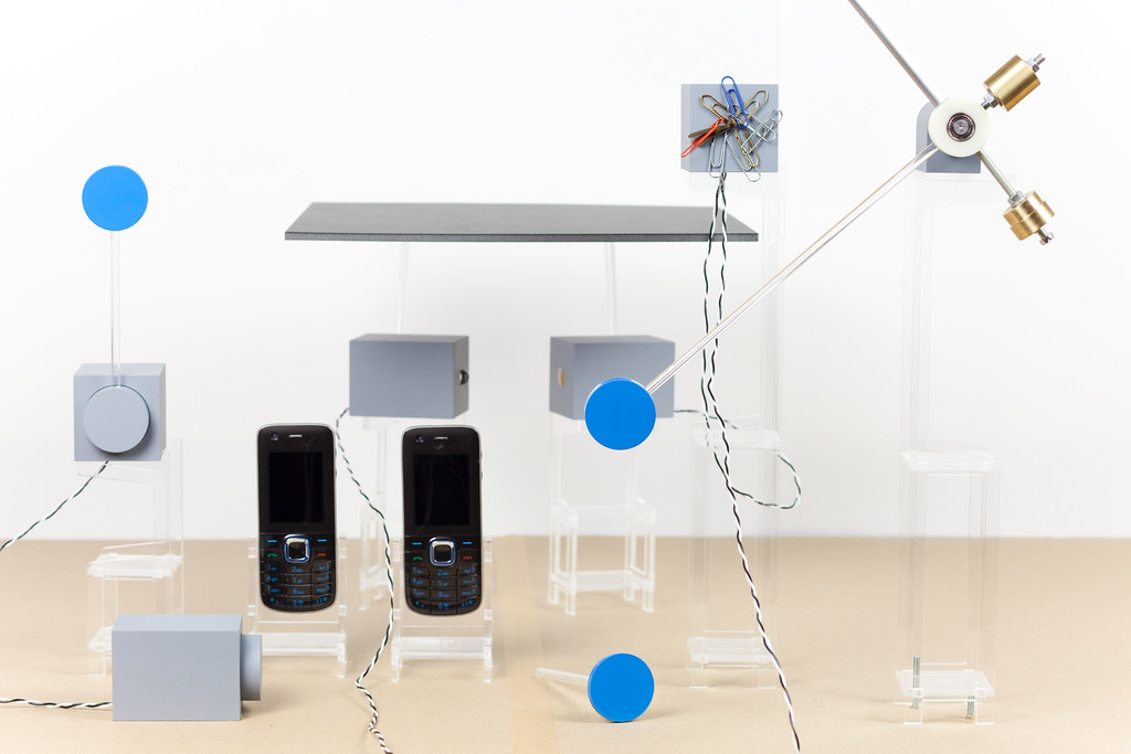 A tabletop assemblage in pale daylight: two Nokia keypad phones on small perspex stands, surrounded by a Fischli-and-Weiss-style mechanical rig of acrylic stages, blue spheres balanced on rods, brass weights on pulleys, and a paperclip-loaded plate. The rig captures the moment two phones touch each other through proximity.