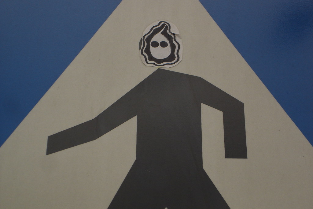 A triangular yield-style traffic sign with a stylised black pedestrian figure. Someone has stuck a small black-and-white sticker over the head: a wavy Munch-mask, two black holes for eyes, like a small ghost face on a sign that has stopped meaning what it once meant.