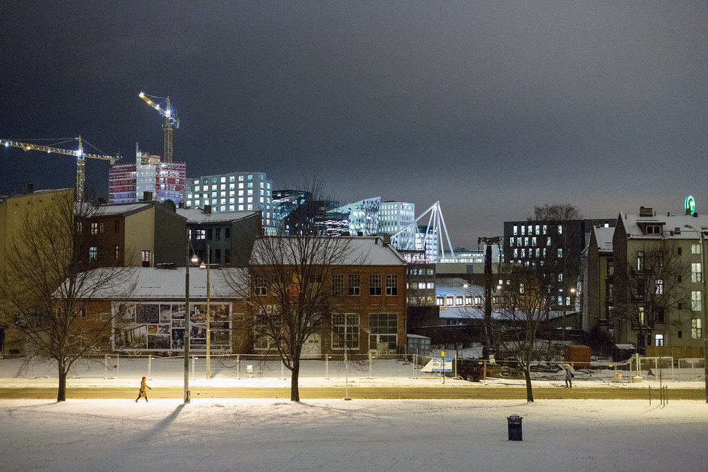 A winter night view across a snowy public plaza in Oslo's Bjørvika quarter. A small figure walks alone across the foreground in a high-vis jacket. Behind them, a long industrial brick warehouse with a mural along its side, and beyond that the lit Barcode buildings and the white slope of the Opera House rising into a heavy grey sky. Two construction cranes lit at the top.