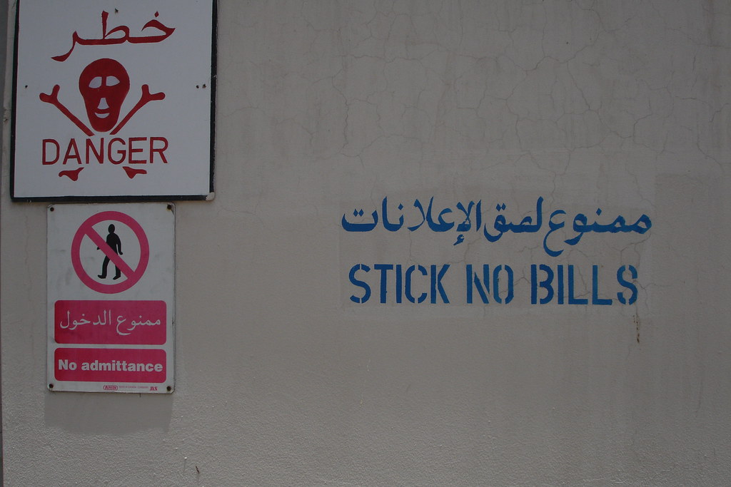 A weathered cream-stucco wall in Dubai. To the upper-left two stacked enamel signs in red: a skull-and-crossbones DANGER warning in Arabic and English, and below it a No Admittance pictogram of a barred figure. To the right, painted directly on the wall in large blue stencilled capitals: STICK NO BILLS, with its Arabic equivalent above.