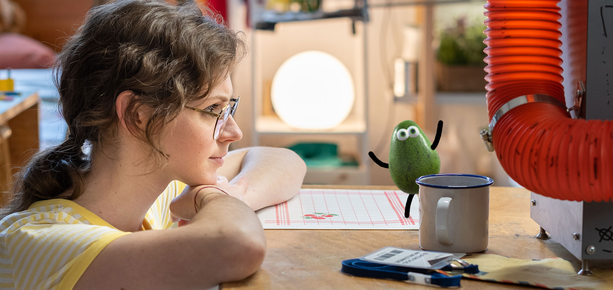 Billie rests her chin on her arm at a workshop bench, watching Avo, the small 3D avocado character, who is balanced on the rim of an enamel mug. An orange ducting and a glowing white sphere are behind them.