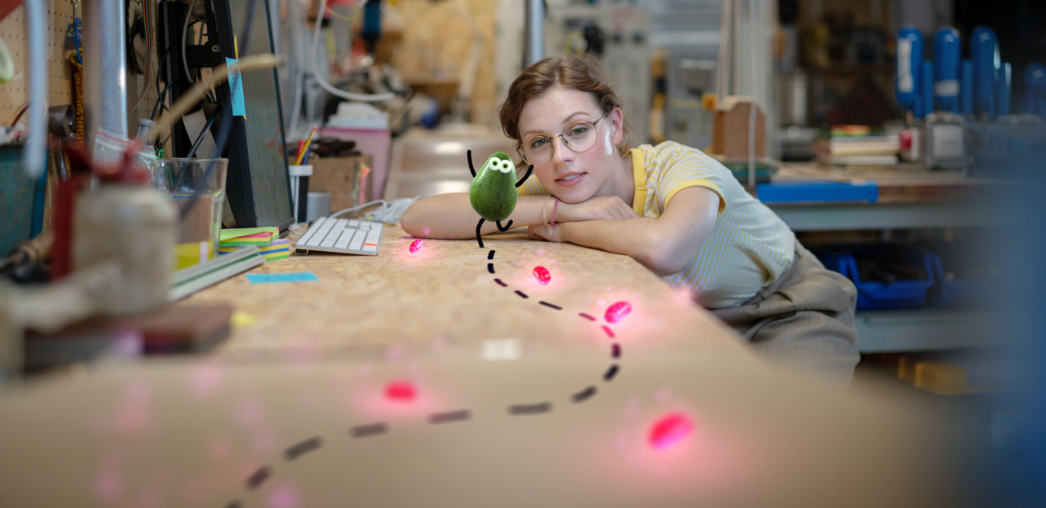 Gameplay still: Avo, a small 3D avocado character with dot eyes and stick limbs, dances on a workshop bench beside Billie, who watches him from across the desk. A dashed path the player has drawn across the wood loops through a trail of glowing pink jellybeans.