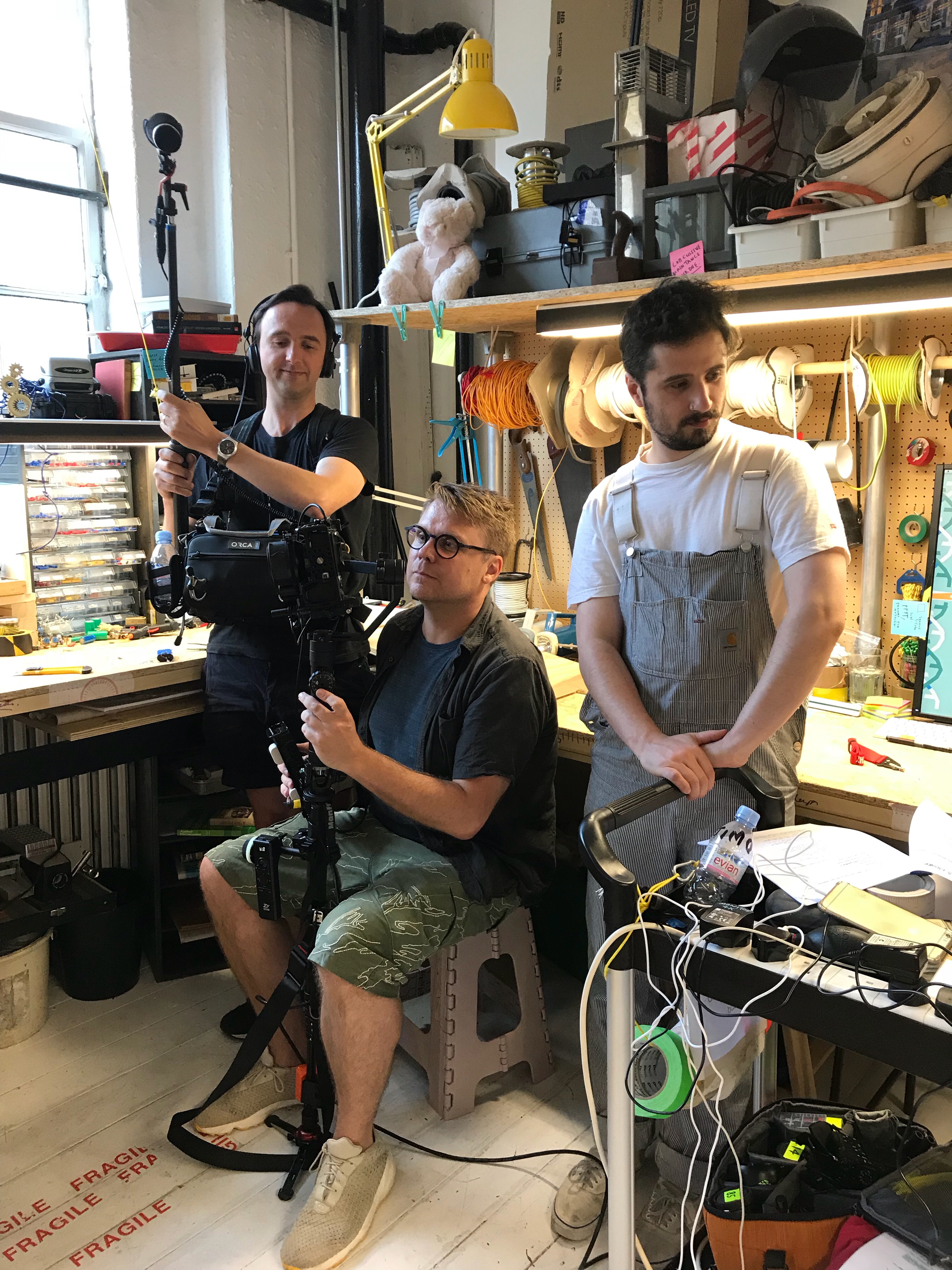 Three of the production crew on set in the studio. One holds a steadicam camera rig, one sits on a stool reviewing a take, one stands with a clapperboard. The studio walls are crowded with shelving full of props and tools.