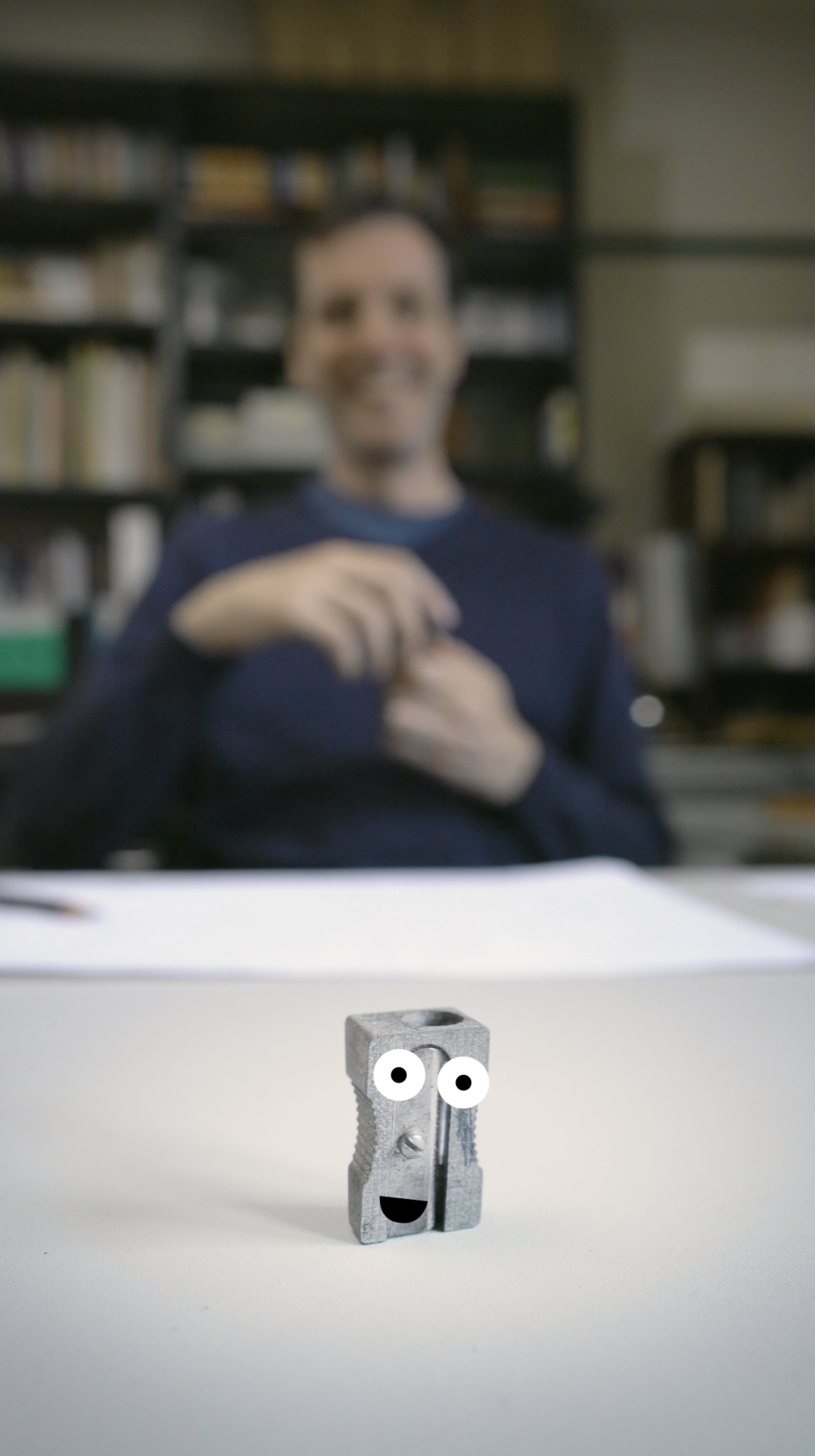 Early prototype: a small metal pencil sharpener with cartoon eyes and a mouth painted on, sitting on a desk. Out of focus behind it, Timo is gesturing at it with one hand.