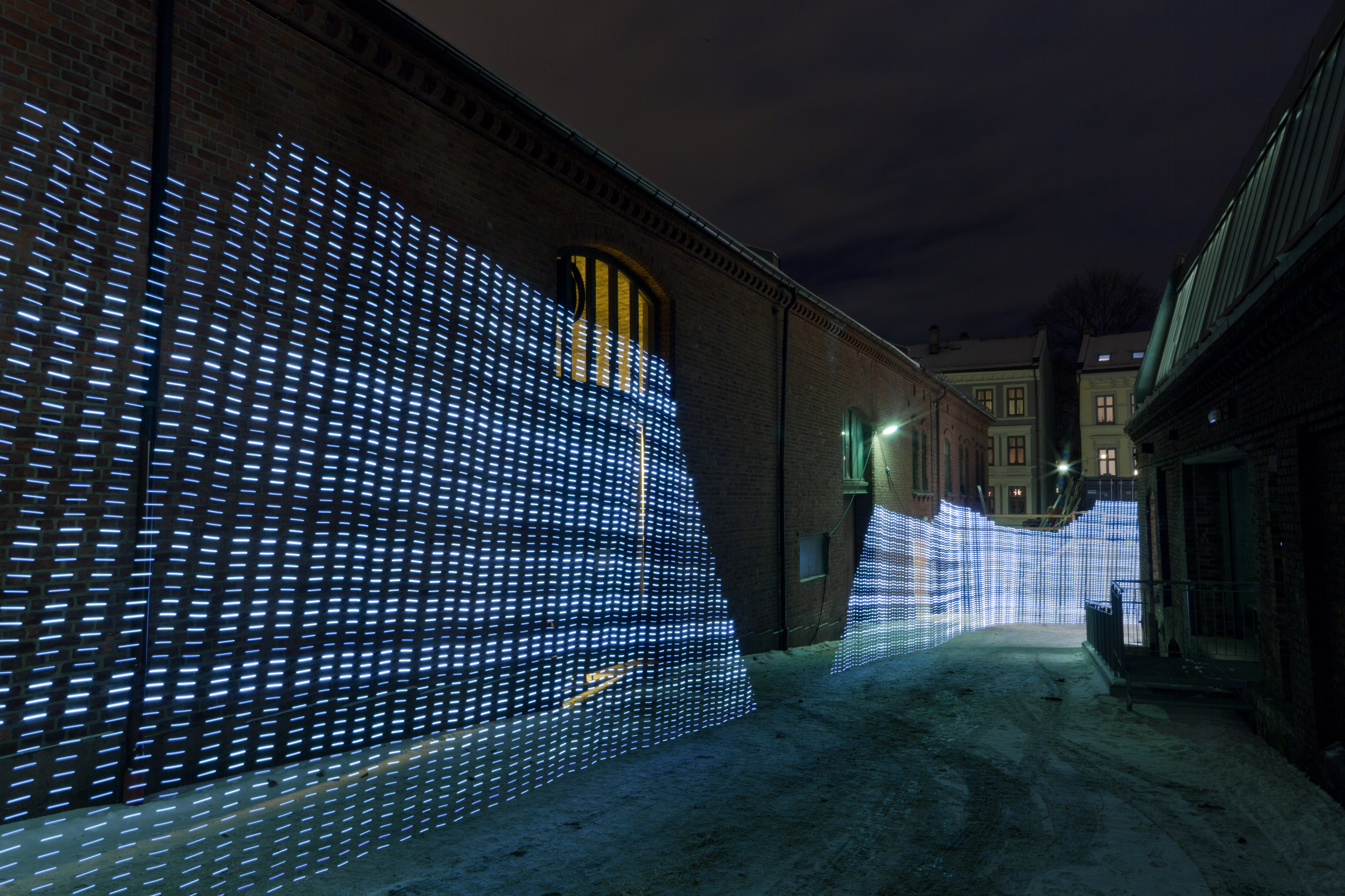 A long-exposure photograph of a courtyard between two brick buildings at night. A continuous wall of pale blue light runs vertically across the scene, partially blocked by the building on the left. The light extends as far as the rod was walked, fading toward the far end of the courtyard.