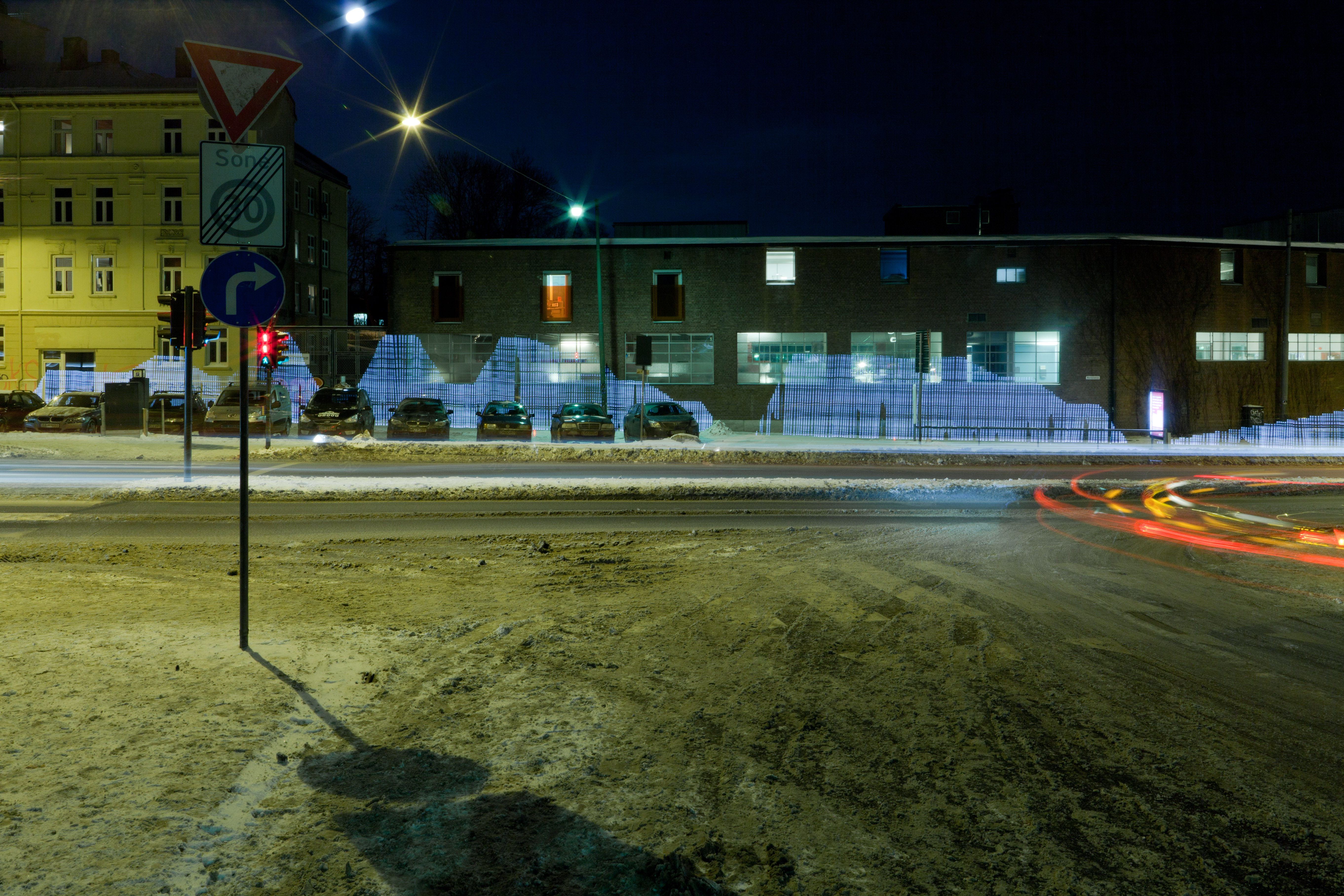 A long-exposure photograph of a street crossing in Oslo at night. A horizontal blue ridge of light hovers in mid-air across the scene, a foot above the ground, the WiFi signal strength rendered as a blue line of pixels. A car has passed, leaving red and white tail-light streaks across the foreground. Snow lies on the pavement.