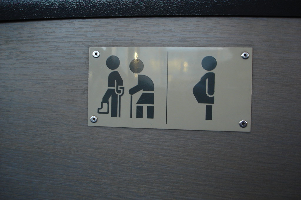 A polished metal sign with three black pictograms on a brushed cream ground: a standing figure with a cane, a seated figure leaning forward with the cane, and a pregnant woman in profile. Mounted to a wood-grain wall panel by four silver studs.