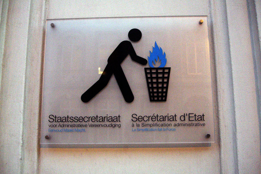 A bilingual Belgian government office sign mounted on a pale wall, reading 'Staatssecretariaat voor Administratieve Vereenvoudiging / Secrétariat d'Etat à la Simplification administrative'. To the right of the text, a black AIGA-style stick figure walks toward a tall waste basket and tosses a document into it; a blue flame rises from the basket. The icon shows an act of disposal-by-burning rendered as the official symbol for administrative simplification.