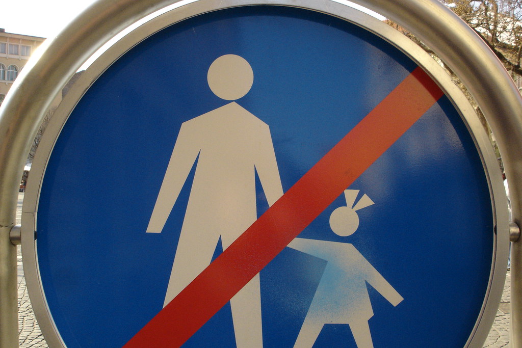A blue circular Slovenian pedestrian-crossing sign showing a white stick figure of a man holding a child's hand against a blue ground. A heavy red diagonal slash, painted directly onto the sign's metal surface, cuts the figures in half — converting the sign's meaning from 'pedestrian crossing' into its inversion: 'no pedestrians'.