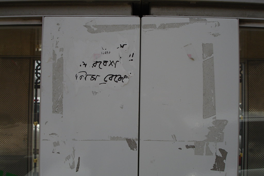 A pair of plain white-tiled door panels in a Paris street, on which dozens of stickers have been pasted and torn off over time. The surface is mottled with adhesive residue, ripped paper edges, faded silver tape, and the worn ghosts of removed posters. Two small remaining stickers carry hand-written script in what appears to be Bengali. The wall behind glows with reflected interior lighting.