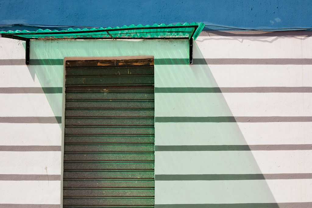 A close-frame photograph of a Tirana facade in Edi Rama's painted programme: a blue band along the upper edge, a green corrugated metal awning, then a series of horizontal bands in white, pale mint, white, and pale blue descending the wall. A green roller-door is set into the centre, casting its diagonal sunlit shadow across the painted stripes. Pure colour-block civic abstraction.