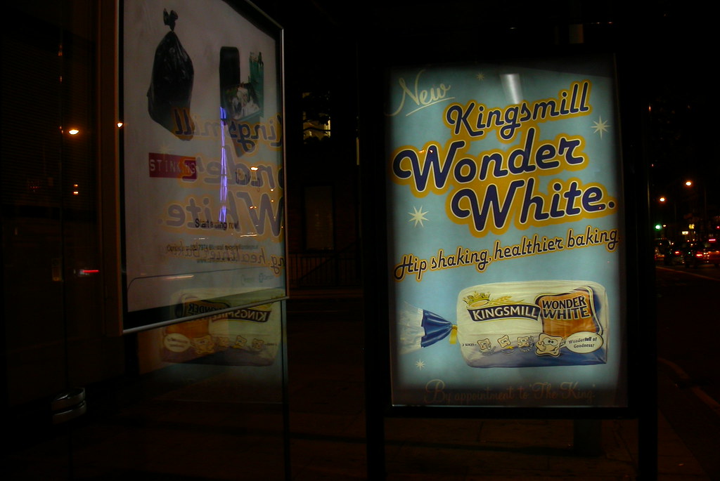 A London bus shelter at night, lit from within. The right-hand panel carries a Kingsmill 'Wonder White' bread advertisement in cheerful 1950s-style typography on a sky-blue ground, with a loaf of sliced white bread and the tagline 'Hip shaking, healthier baking'. The left-hand panel reflects a torn black bin-bag stuck to its surface in approximately the same shape and position as the bread loaf — a quietly devastating subversion that uses the panel itself as the canvas.