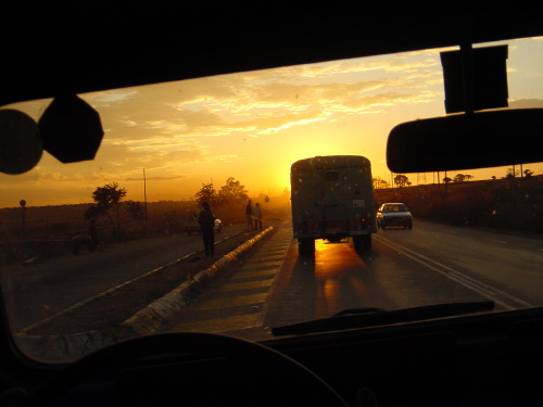 A Zimbabwean road at sunset, a battered minibus ahead throwing golden dust, two pedestrians silhouetted at the verge.