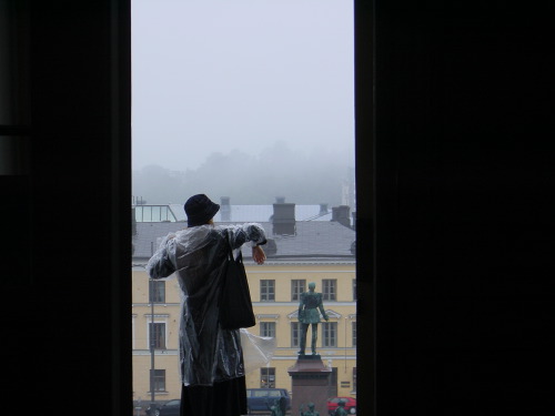 Looking out through a Helsinki window at a hooded figure in a translucent rain poncho leaning over the parapet, a small bronze equestrian statue and pale-yellow Helsinki buildings dissolved in heavy fog beyond, the dark window frame at left and right.