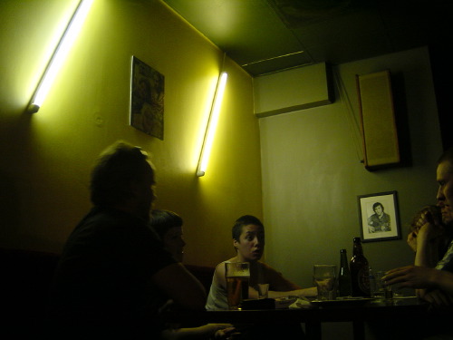 A late-night Helsinki bar interior, ochre walls under cool fluorescent strip lights, three patrons seated around a small table with beer bottles, two framed pictures on the wall, a fourth diner half-cropped in shadow at the right edge.