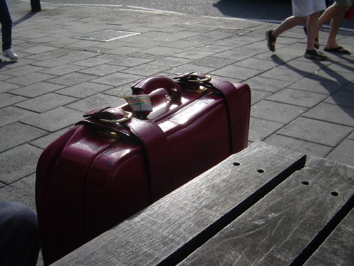 A red leather suitcase resting on a wooden public bench in slanted late-afternoon sunlight, the long shadow of the bench falling across the cobbled pavement. The legs of two pedestrians are visible mid-stride at the top of the frame.