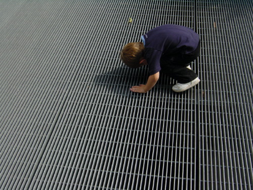 A child in dark blue clothing crouched on a metal grating walkway, head down, looking through the grid at whatever lies beneath. The grating fills almost the whole frame as a strict orthogonal lattice.