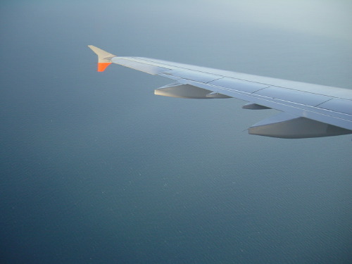 An aircraft wing photographed through a passenger window: the wing-tip with its small orange winglet against a calm grey sea below, the engine cowling visible at the right edge.