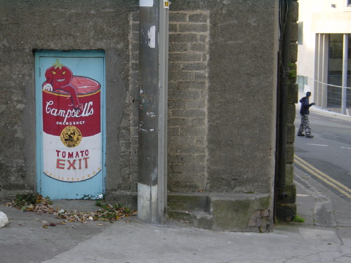 A side-street corner in Wexford, Ireland: a Campbell's-Soup-pastiche poster reading 'Tomato Exit' painted onto a doorway in a stone wall. Beyond the corner, a lone figure walks down a sunlit side-street between brick and limewashed buildings.