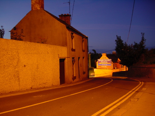 A small Wicklow village street at deep blue twilight: a low whitewashed cottage on the left, a 'Murphy's Sport Pub' lit sign visible further along, sodium lamps casting warm pools of light onto wet asphalt and yellow road markings.