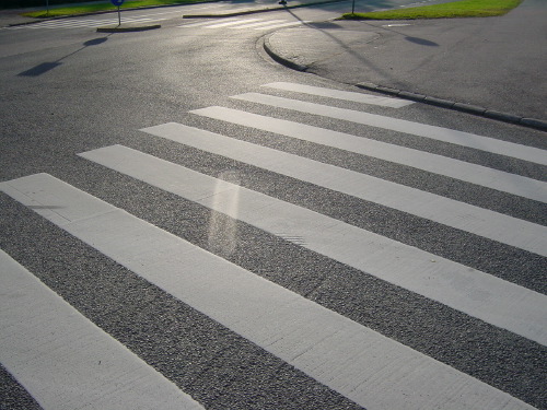 A wide pedestrian crossing on an empty Espoo street in raking late-afternoon sun, the painted white stripes casting long shadows on grey asphalt. The geometry fills almost the whole frame.