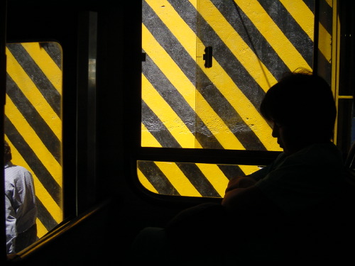 Inside a Buenos Aires train carriage: a service door painted in heavy yellow-and-black diagonal hazard stripes, almost filling the frame. A passenger's silhouetted head and shoulder dominate the right side; another passenger is faintly visible through the door's small window.