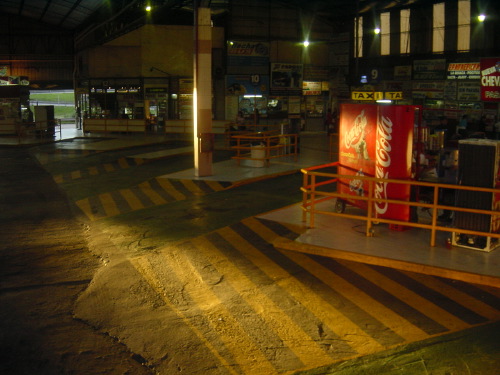 An empty Buenos Aires bus terminal at dusk: yellow-and-grey diagonal floor tiles, a red Coca-Cola fridge, sodium lights overhead, signage receding into the back of the hall.