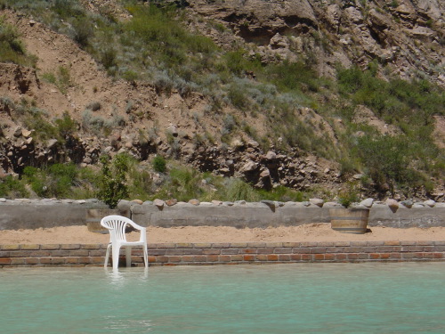 A pale-turquoise swimming pool fed from a rocky desert hillside near Mendoza, a single white plastic chair half-submerged at the edge, brick paving and scrubland beyond.