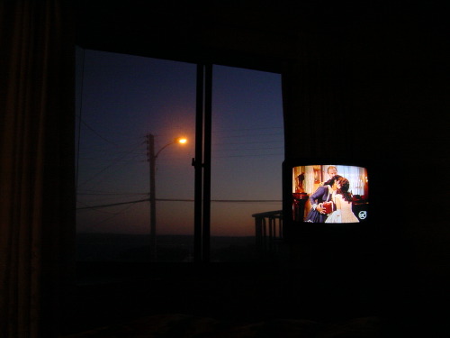 A motel room in Caldera, Chile, at dusk: a small CRT television to the right showing a black-and-white film scene of two figures embracing, and a window to the left showing a streetlamp glowing orange against a deep blue evening sky.
