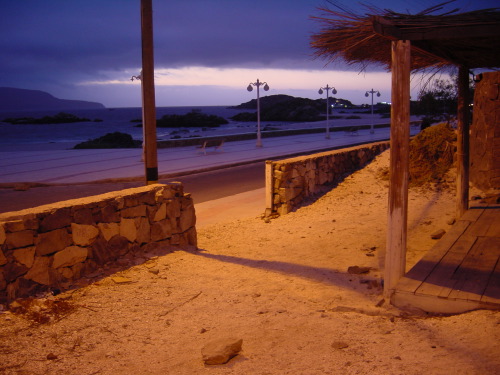 A stone-walled boardwalk at dusk in Bahía Inglesa: rusted-orange sodium light pooling on the sand in the foreground, the dark headlands and empty beach beyond, a small line of seafront streetlamps, deep blue clouds above.