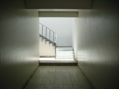 A bright modernist concrete corridor in Calama, looking through a doorway onto a flight of stairs ascending behind tubular-steel railings. The walls are white, the floor grey-blue tile, the space almost entirely empty.