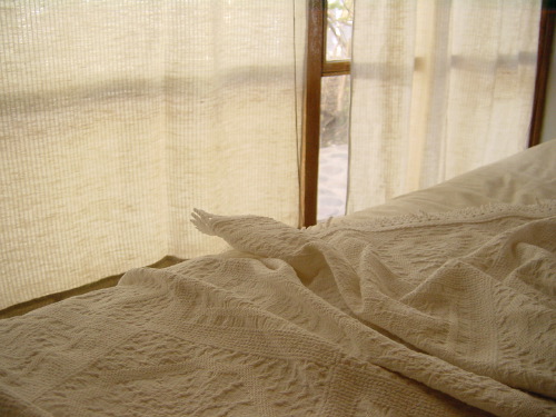 A bedroom interior in San Pedro de Atacama: a heavy cream woven bedspread, soft thin curtains catching daylight at a window with a wooden frame, a small bright slice of sky and stone visible beyond.