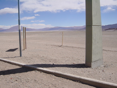 A roadside rest stop on the Andean altiplano between San Pedro and Jujuy: two metal poles framing a flat brown plain, distant blue mountain ranges, scattered cumulus cloud, a concrete-edged road in the foreground. Brilliant midday light.
