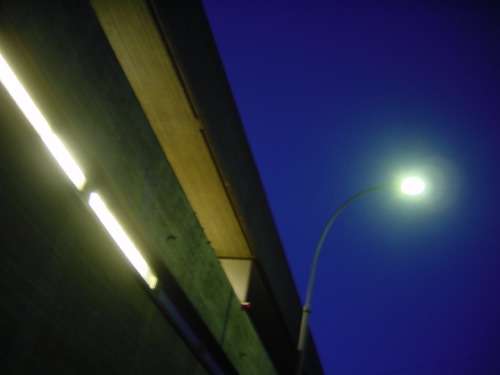 A concrete underpass at deep blue twilight, a strip of fluorescent light running along its eaves, a single curved lamppost on the right with a bright sodium head against a darkening sky.