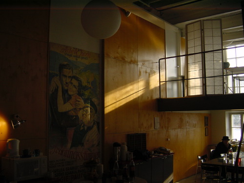 A loft-converted café in Helsinki, golden afternoon light pouring in from tall industrial windows on the right, a large vintage Pierre Brasseur film poster on the left wall, a small lamp on the bar in the foreground, a lone bowler-hatted figure seated at a table at the far right.