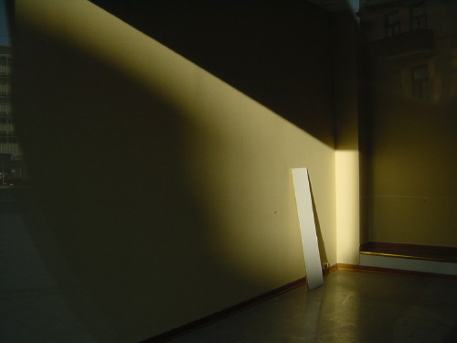 An Oslo apartment hallway at dusk: a wedge of warm sunlight cutting diagonally across a yellow-cream wall from an off-frame window, a thin white door panel propped vertically against the wall, the brick fronting of a building across the street visible through the doorway.