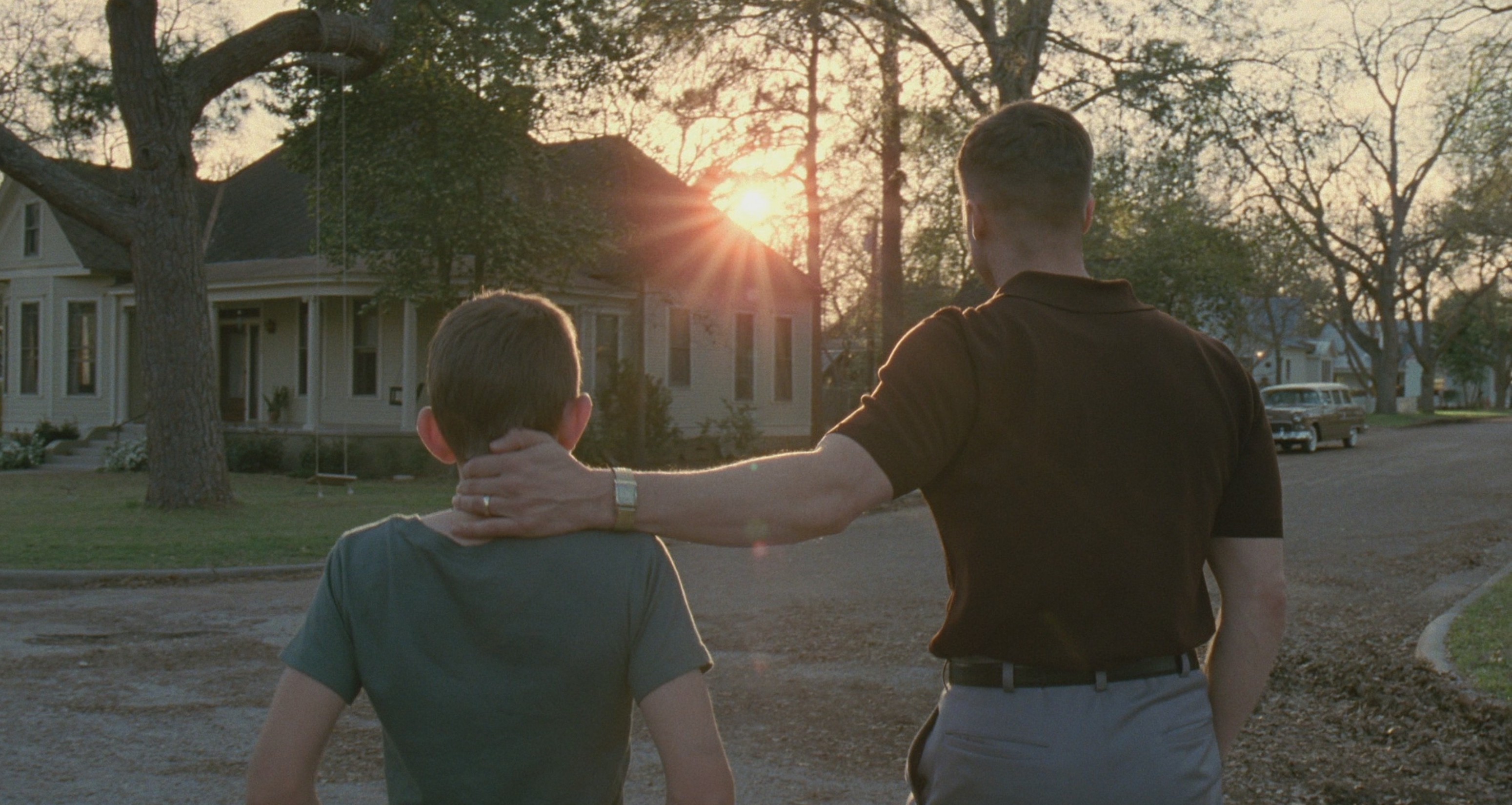 A father and his son walk away from camera down a quiet neighbourhood street at sunset. The father has his hand on the back of the boy's neck. A 1950s house and a wide oak frame the scene; sun-flare comes through the trees ahead. From The Tree of Life, Terrence Malick, 2011.