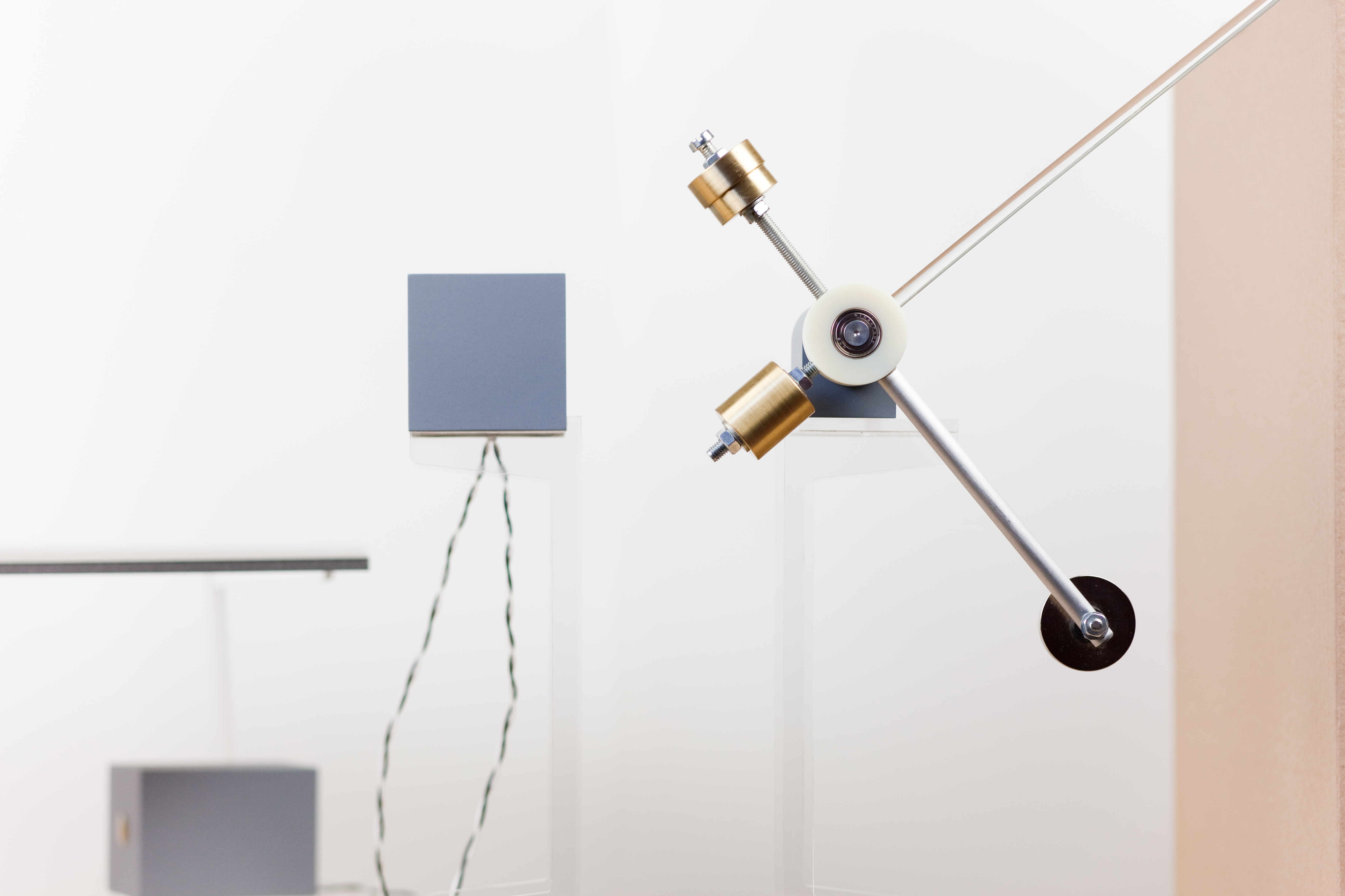 A close-up of one of the small mechanisms in the rig: a precise brass-and-aluminium balance arm pivoting on a white plastic wheel, with two cylindrical brass weights on a threaded shaft. The arm extends out of frame at top right and supports a small black disc bottom right. Behind, a grey block on an acrylic stand and a wire dropping from a second block.