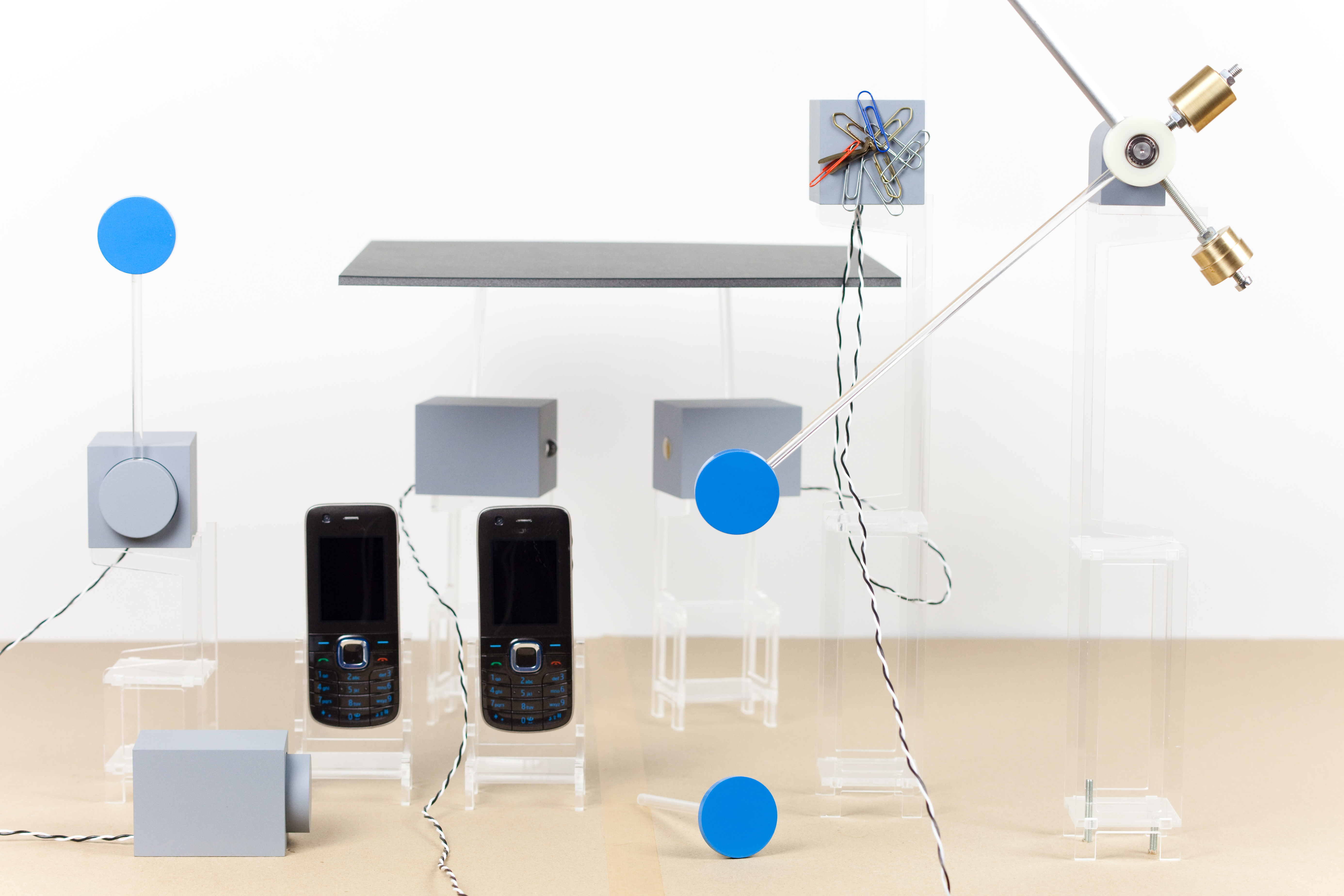 A wide shot of the Nearness rig: a tabletop scene of acrylic stands, grey painted blocks, blue circles on rods, two old Nokia phones in the centre, an articulated arm with brass weights, a paperclip jumble, and slender wires connecting components. Soft daylight. The composition reads as the kind of intricate kinetic stage a chain reaction could run across.