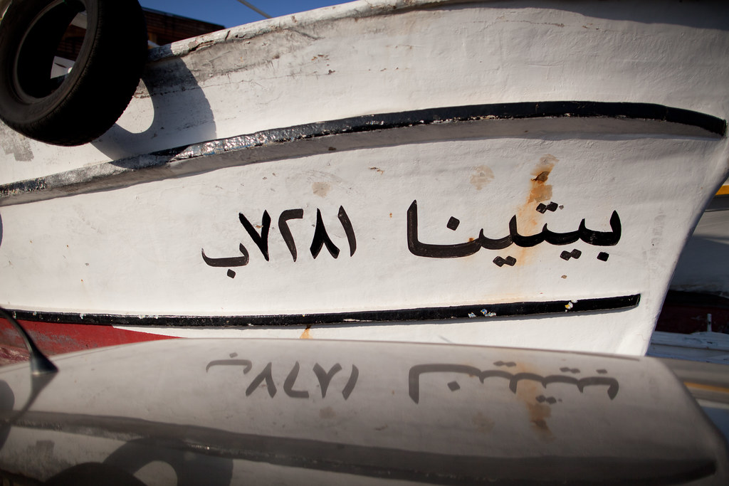 A close, sun-flat photograph of the white-painted bow of a wooden fishing boat. Black hand-painted Arabic script reads بيتينا and a registration number 7281ب. A black tyre fender hangs over the bow at the upper-left, throwing soft shadow. The reflection of the same script appears upside-down on the polished black surface of an adjacent vessel below.