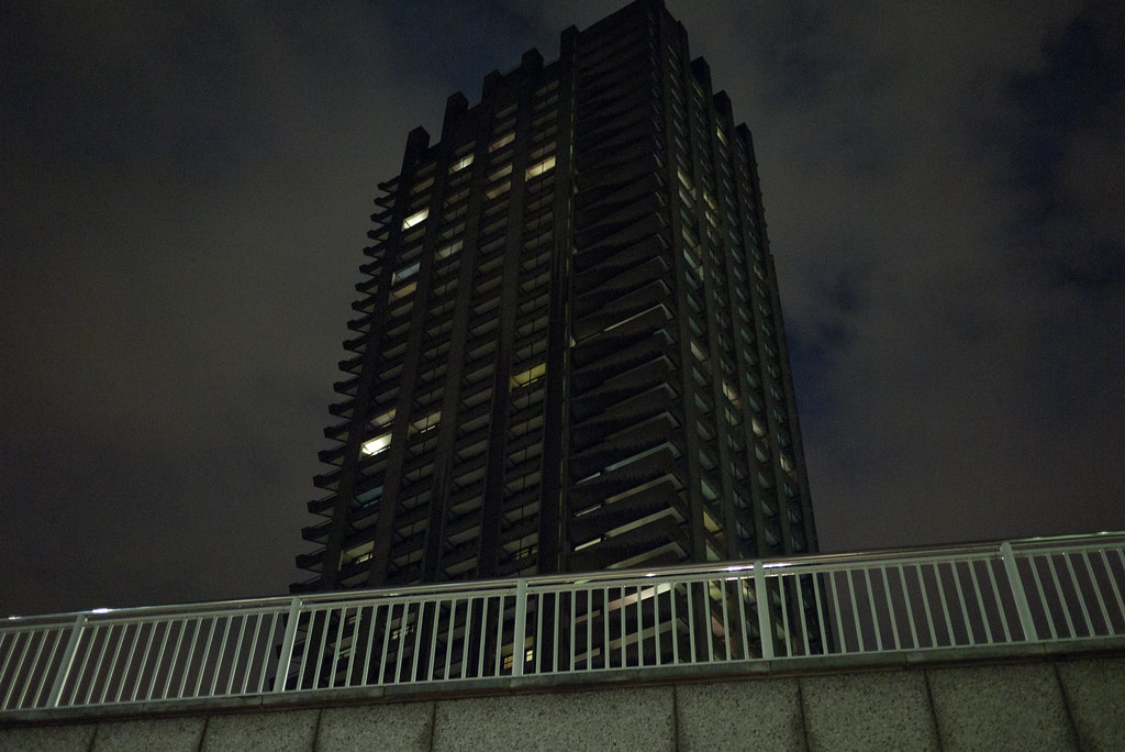 A vertical view looking up at a Barbican tower at night. The brutalist concrete-and-iron facade rises against a hazy black-grey sky, the deep balcony rails reading like comb teeth, only a few rooms lit in soft warm yellow. A railing along a walkway in the foreground reflects the city's ambient light; a stone retaining wall fills the bottom edge of the frame.