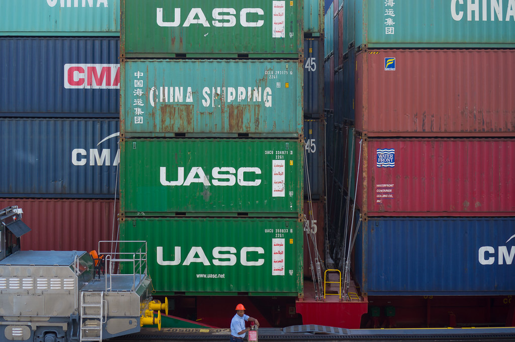 A telephoto frame of a port container yard. Stacks of shipping containers fill the frame in horizontal bands of saturated colour: deep navy, red CMA CGM units, vivid green UASC units stencilled CHINA SHIPPING in white, a thin stripe of yellow industrial machinery along the bottom. A single small worker in a red hard-hat stands at the base of the stack, the only human in a wall of trade.