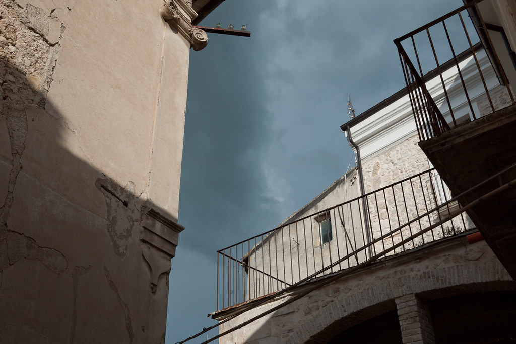 A view from a narrow Italian alley looking up at a stormy blue-grey sky. To the left, a peeling cream-stucco wall and a small cornice from which two small lamps are suspended on an iron arm. To the right, a partially balconied Italian house with rusty wrought-iron railings, white-painted shutters, and a small turquoise window catching grey light. The composition reads as architecture-from-below as a single tall vertical breath of sky.