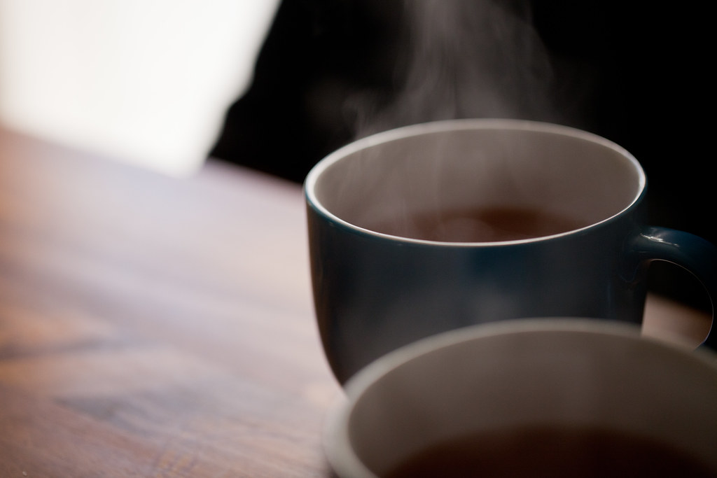 A shallow-depth-of-field morning still life: two ceramic teacups on a wooden table, steam rising from each cup into the shadow of the room above. The closer cup is dark blue, the further cup white, both half-filled with dark tea. Bright window light blows out the left edge of the frame; the rest of the scene is muted in cool greys and warm browns.